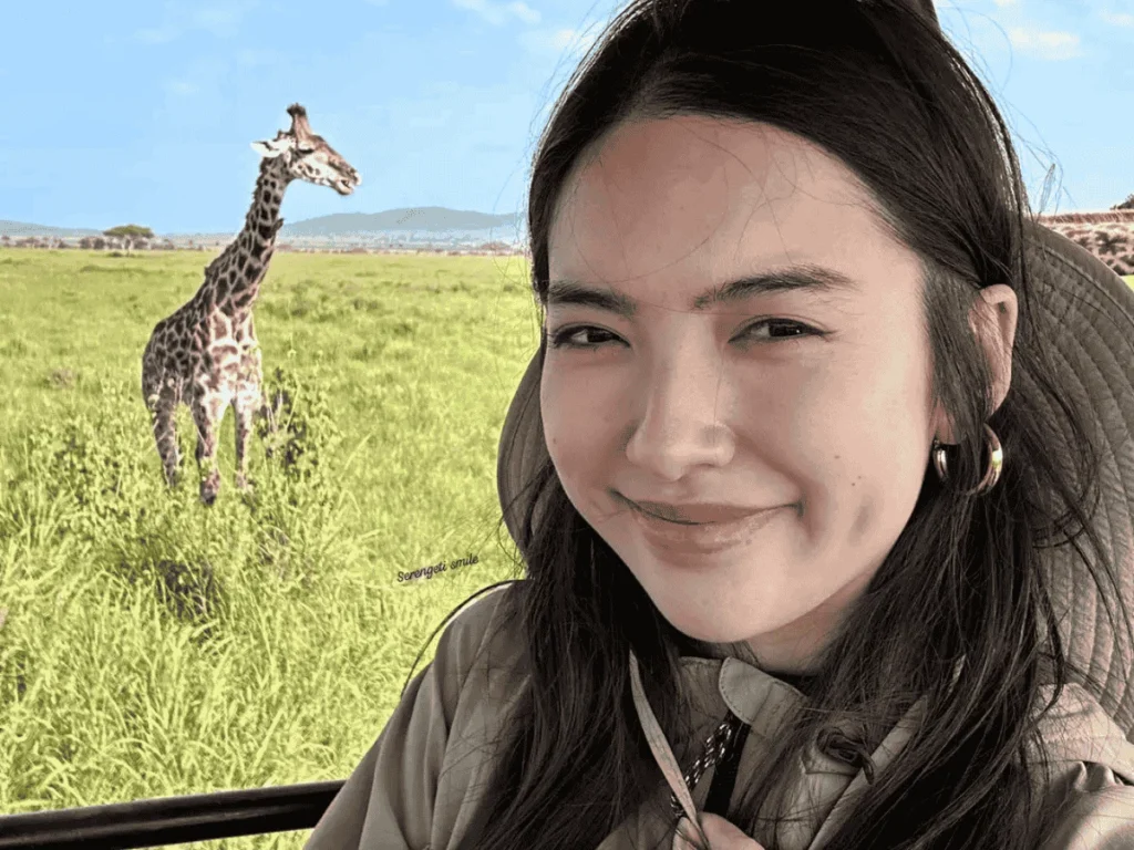 A young woman enjoys a wildlife safari in Lake Manyara National Park with the Maus Safari Company. A giraffe can be seen in the background.