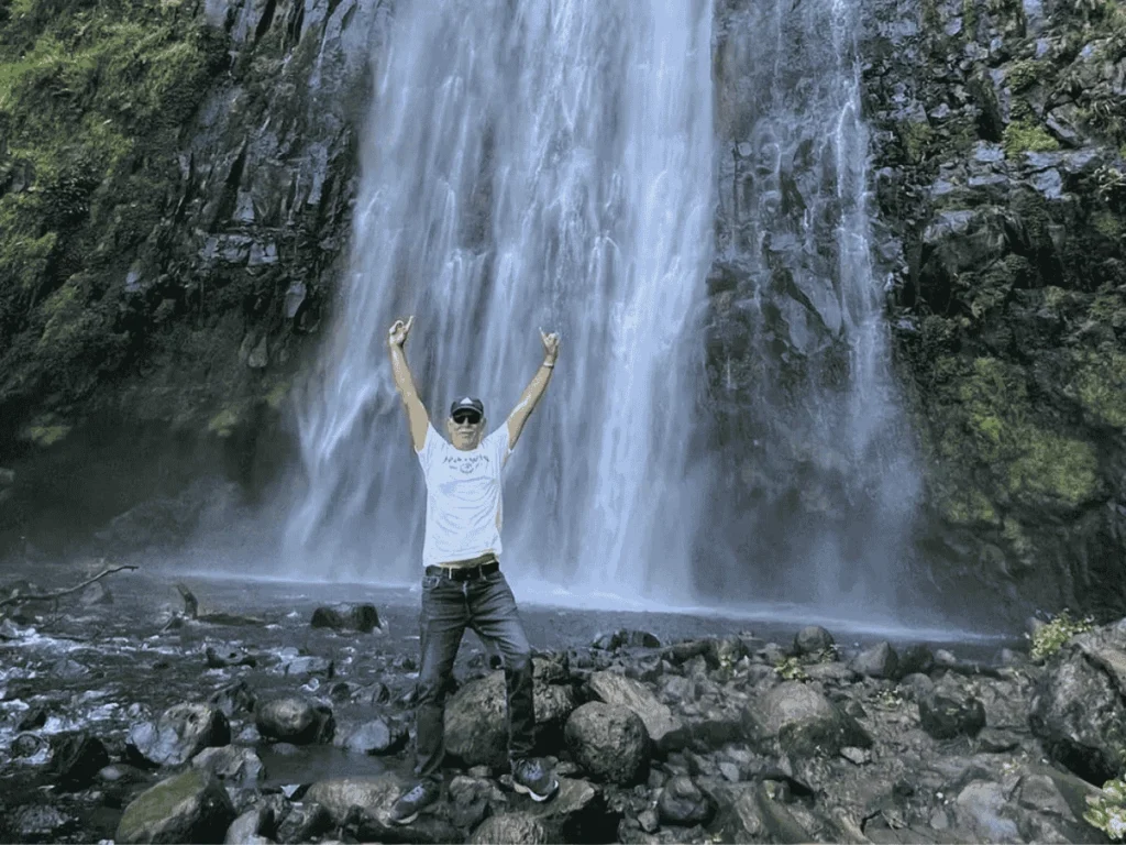 A man stands in front of a waterfall with his arms outstretched, surrounded by spraying water and lush greenery. Day Trip Materuni Waterfalls.