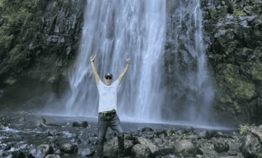 A man stands in front of a waterfall with his arms outstretched, surrounded by spraying water and lush greenery. Day Trip Materuni Waterfalls.