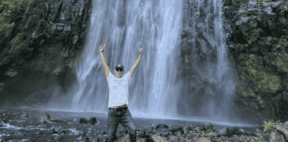 A man stands in front of a waterfall with his arms outstretched, surrounded by spraying water and lush greenery. Day Trip Materuni Waterfalls.