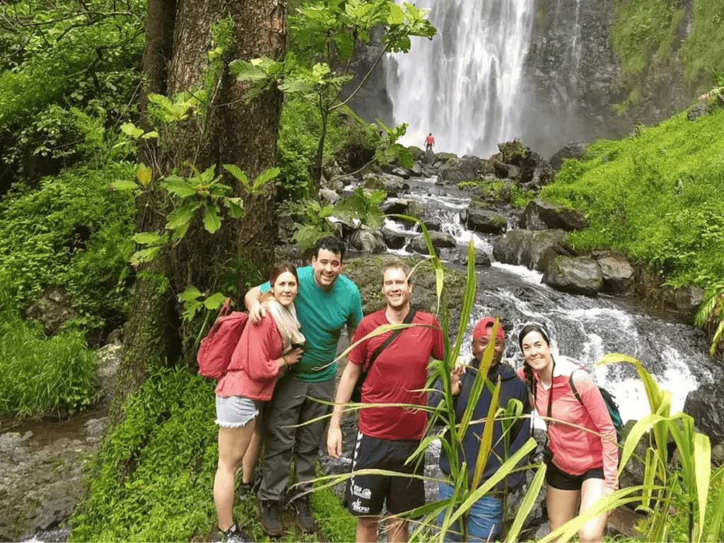 A group of smiling men and women pose in front of an impressive waterfall. Materuni Waterfalls.